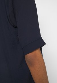 Camisetta blu navy a maniche corte con risvolto, caratterizzata da una texture morbida e liscia e da una vestibilità rilassata, che mostra parte di un braccio accanto ad essa.