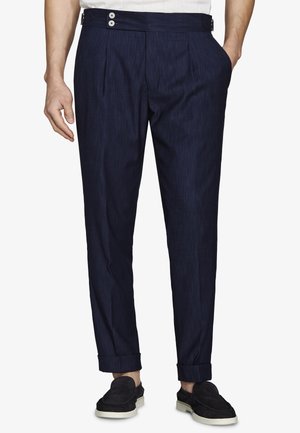 Męskie granatowe dopasowane spodnie z bocznymi guziczkami do regulacji, z mankietami na nogawkach, noszone z czarnymi wsuwanymi butami, ręce swobodnie ułożone wzdłuż ciała.