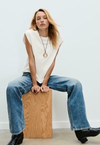 T-shirt crème oversize à manches courtes, associé à un jean large bleu clair. Le mannequin est assis sur une boîte en bois, portant des bottines noires.