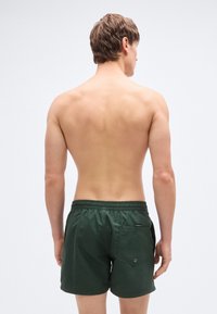 Σκούρα πράσινα μαγιό σορτς με ελαστική μέση, πίσω τσέπη με μεταλλική τ ρύπα και απαλό υφασμάτινο υφή. Θέα από πίσω.