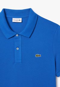 Polo azul hecho de tela texturizada. Presenta un cuello doblado, dos botones y un logotipo de cocodrilo bordado en verde en el lado izquierdo del pecho.