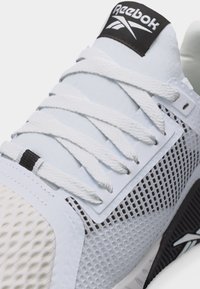 Λευκό αθλητικό παπούτσι Reebok με υφασμάτινη πλέξη, λευκά κορδόνια και μαύρο λογότυπο στη γλώσσα και στην πλευρά, τοποθετημένο λοξά για να δείξει λεπτομέρειες από το πάνω μέρος και την πλευρά.