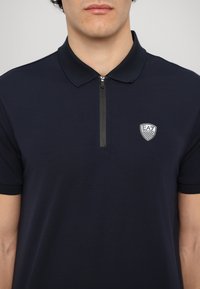 Námornícky modrá polo tričko so zipsom, krátkymi rukávmi a malým bielym logom na ľavej strane. Vyrobené z hladkej, ľahkej tkaniny.