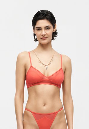 Kobieta o krótkich ciemnych włosach, ubrana w koralowo-czerwony biustonosz i dopasowane majtki z wysokim stanem, stojąca na tle jednolitego jasnego tła.