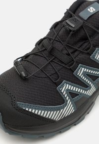 Zapato deportivo negro con parte superior de tela texturizada, sistema de cordones elásticos y un acento gris con patrón. Suela de goma duradera para mayor tracción.