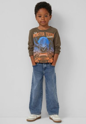 Braunes Langarmhemd mit buntem Monstertruck-Design, kombiniert mit weit geschnittenen blauen Jeans und weißen Sneakers mit braunen Sohlen.