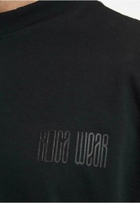 Black cotton T-shirt with a ribbed neckline, featuring a raised black 'ROCA WEAR' logo on the front. Smooth texture and minimalist design.