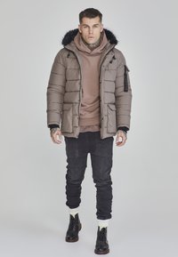 Pufferjakke i dæmpet brun med en futteret hætte, iført over en beige hoodie, parret med sorte jeans og støvler.