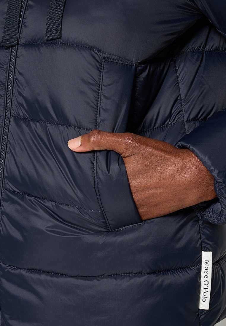 Navy Puffer-Jacke mit gesteppten Abschnitten, ausgestattet mit einer Seitentasche und einem Etikett. Die Textur wirkt glatt und leicht glänzend.