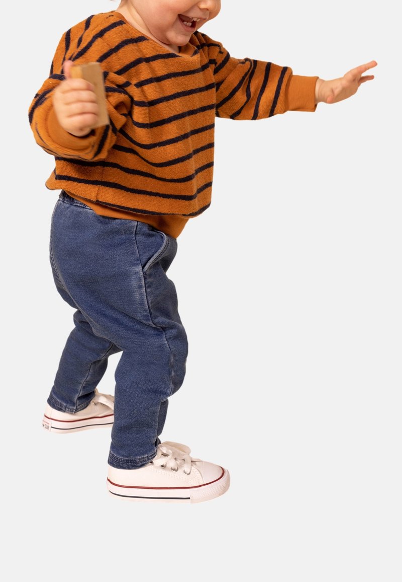 Enfant portant un pull rayé orange et un pantalon en denim bleu, avec des baskets blanches à accents rouges, tenant un petit objet tout en marchant.