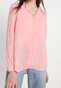 Rosa, lätt blus med V-ringning och långa ärmar. Har en slät textur och en något rundad nederkant, samt passar bra med blå denim.