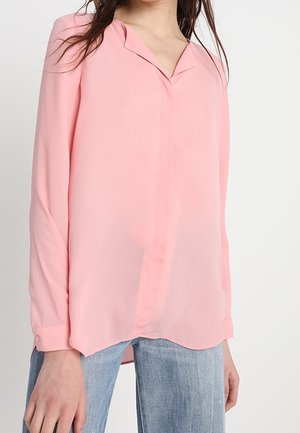 Blusa leggera rosa con scollo a V e maniche lunghe. Presenta una texture liscia e un orlo leggermente curvo, abbinata a jeans blu.