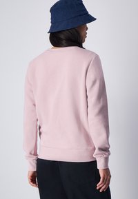 Felpa rosa chiaro con scollo rotondo e maniche lunghe. Tessuto morbido. Indossata con un cappello a secchiello blu navy, mostrando la vista posteriore.