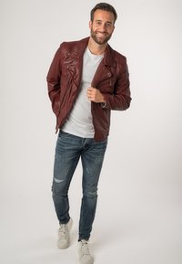 Giacca biker in pelle rossa con dettagli in argento, indossata sopra una maglietta grigia, abbinata a jeans blu sdruciti e sneakers bianche.