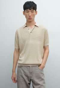 Strikket beige polo med struktureret mønster, korte ærmer og en lys farvet krave. Kombineret med lysegrå jeans.