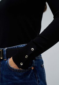 Haut à manches longues noir avec des poignets côtelés et des accents de boutons argentés, porté avec un jean en denim bleu, présentant un détail de poche.