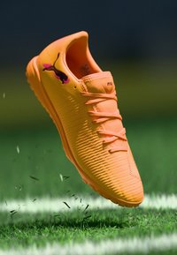 Oranger Sportschuh mit strukturiertem Design, Schnürsenkeln und einem pinkfarbenen Logo. Schwebt über grünem Gras mit verstreutem Schmutz.