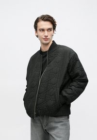 Νεαρός με κυματιστά μαλλιά, φοράει μαύρο μπουφάν bomber με σχέδια πάνω από μαύρο πουκάμισο και γκρι τζιν, τα χέρια στις τσέπες του μπουφάν, κοιτάζει πλάγια.