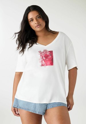 Giovane donna con lunghi capelli scuri che indossa una T-shirt bianca con scollo a V e stampa floreale rosa, abbinata a una gonna a portafoglio in denim blu su sfondo bianco.