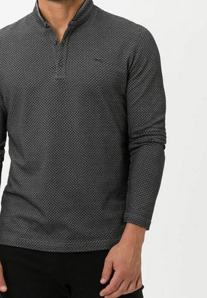 Mand iført en mørkegrå tekstureret langærmet poloshirt med en tre-knaps lukning og sorte bukser, stående foran en ensfarvet baggrund.