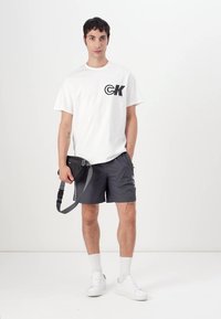Balta medvilninė marškinėliai su juodu "CK" logotipu, suporuoti su pilkais šortais, baltais kojiniais ir baltais sportbačiais. Modelis laiko juodą krepšį per petį.