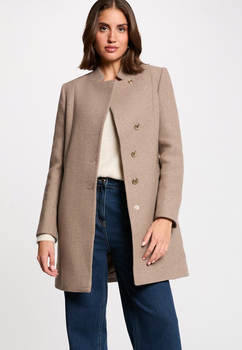 Abrigo de mezcla de lana beige con corte recto, que presenta un cuello alto, cinco botones en tono dorado y bolsillos laterales, combinado con jeans de pierna ancha.