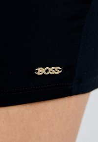 Juodi prigludę šortai iš tamprios medžiagos su auksiniu "BOSS" logotipo ženkleliu, prisisiuvtu prie krašto. Lygus paviršius ir jokių matomų raštų.