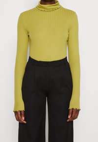 Vihreä ribattua turtleneck-neuletta, jossa on röyhelökaulus ja pitkät hihat, yhdistetty mustiin korkea vyötäröisiin leveälahkeisiin housuihin.