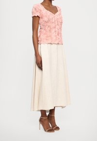 Haut à manches courtes texturé floral rose associé à une jupe beige longueur cheville. Il présente un design en V et des sandales à talons simples avec des lanières.