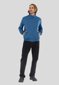 Leichte blaue Jacke mit Reißverschluss, Stehkragen und elastischen Bündchen. Kombiniert mit dunklen Hosen und grauen Schuhen mit strukturierten Sohlen.