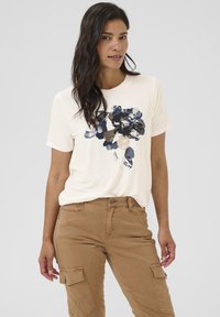 Cremefarbenes T-Shirt mit kurzen Ärmeln und einem navyfarbenen Blumenprint in der Mitte, kombiniert mit khakifarbenen Cargo-Hosen mit Seitentaschen. Weicher Stoff, lockere Passform.