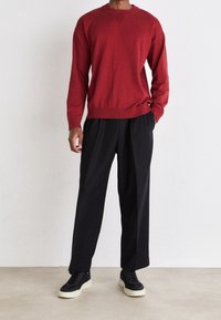 Roter Strickpullover mit rundem Halsausschnitt und gerippten Bündchen, kombiniert mit lockeren schwarzen Hosen und schwarzen Turnschuhen mit cremefarbenen Sohlen.
