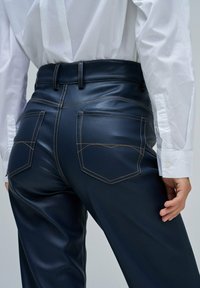 Pantalon en simili cuir bleu marine avec des coutures jaunes, doté de deux poches arrière et d'une coupe ajustée, associé à une chemise blanche.