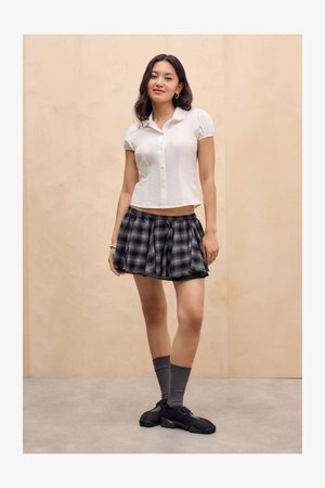 Jeune femme se tenant à l'intérieur, portant un blouse blanche à manches courtes, une jupe à carreaux noire et grise, des chaussettes grises et des baskets noires.