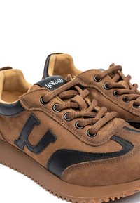 Brown suede sneakers with black accents, featuring a raised logo, round laces, and a textured rubber sole.