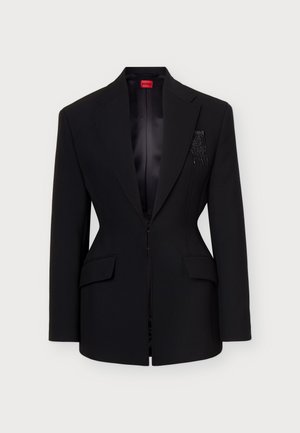 Femme de face portant un blazer noir ajusté avec des embellissements, un pantalon noir évasé et un haut blanc col montant sur un fond clair uni.