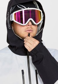 Hvid hættejakke med sorte detaljer, lynlåslukning foran og justerbare manchetter. Bæres med store hvide og pink spejlede skibriller.