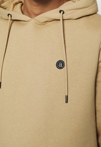 Felpa beige in cotone con cappuccio e lacci, caratterizzata da un logo nero circolare sul petto e terminali neri sui lacci. Tessuto morbido.