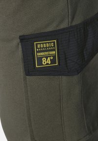 Olivgrüne Stoffhose mit marineblauen Details. Verfügt über ein rechteckiges schwarz-gelbes Etikett mit der Aufschrift „NORDIC WOODLANDS VANDERSTORM 84°.“