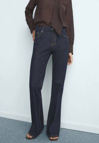 Person trägt dunkelblaue Jeans und ein braunes Langarmhemd, steht barfuß auf einem grauen Teppich vor einer weißen Wand.