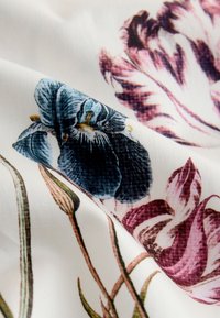Vitt tyg med stora detaljerade blå och lila blommor och gröna stjälkar tryckta på det, visat med mjuka veck.