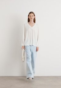 Witte blouse met lange mouwen en V-hals, met een trekkoord, gecombineerd met lichtblauwe wijde jeans. Model houdt een lichtgekleurde tas vast.