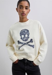 Sudadera crema con cuello redondo. Presenta un gráfico de calavera y huesos cruzados en color azul marino. Textura suave y corte relajado, con puños de canalé.