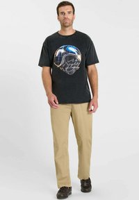 Zwart grafisch t-shirt met een helmontwerp, gecombineerd met beige broek. Model draagt donkere schoenen. Simpele, casual kleding weergegeven tegen een neutrale achtergrond.