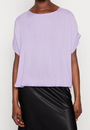 Kvinde iført en løs lavendelfarvet t-shirt med korte ærmer kombineret med et sort satin skørt, vist fra skuldre til hofter mod en ensfarvet baggrund.