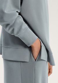 Sivý sveter s hladkým povrchom, ktorý má bočnú kapsu a rebrovaný detail na leme. Voľný strih s dlhými rukávmi.