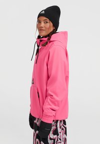 Felroze softshell jas met een capuchon, een ritszak aan de voorkant en elastische manchetten. Gecombineerd met zwarte handschoenen en patroonbroek.