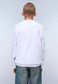 Hvit hettegenser med lange ermer og ribbestrikkede mansjetter, kombinert med lyseblå denimjeans. Enkel design, ingen synlige mønstre eller grafikk.
