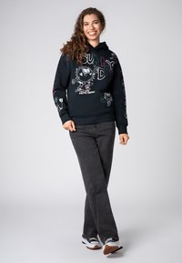 Zwarte hoodie met kleurrijke grafische print van een cartoonfiguur. In combinatie met donkergrijze jeans. Wit en zwarte sneakers.