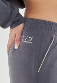 Pantaloni jogger grigi con vita elastica, adornati con logo EA7 e dettagli in cristallo lungo la tasca. Texture morbida e liscia.